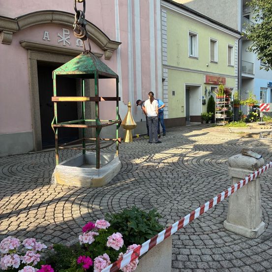 Zwei Personen befinden sich in der Nähe eines grünen Metallobjekts mit Blumen und Pflanzen darum. Es gibt ein Gebäude mit einem Schild über der Tür.