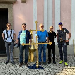 Fünf Männer stehen vor einem Gebäude und halten ein großes goldenes Kreuz. Sie sind in Freizeitkleidung gekleidet und scheinen für ein Foto zu posieren.