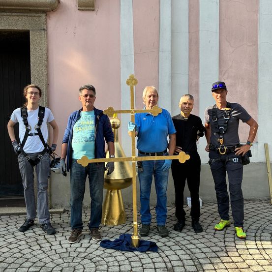 Fünf Männer stehen vor einem Gebäude und halten ein großes goldenes Kreuz. Sie sind in Freizeitkleidung gekleidet und scheinen für ein Foto zu posieren.