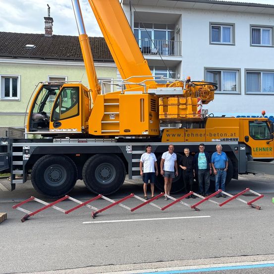 Fünf Männer posieren vor einem großen gelben Liebherr-Kran mit ausgefahrenem Ausleger, der auf einer Straße vor einem mehrstöckigen Gebäude positioniert ist.