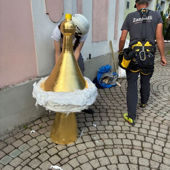 Eine Person mit Helm wickelt einen goldenen Kegel mit einer weißen Abdeckung ein. Eine weitere Person geht neben ihm.