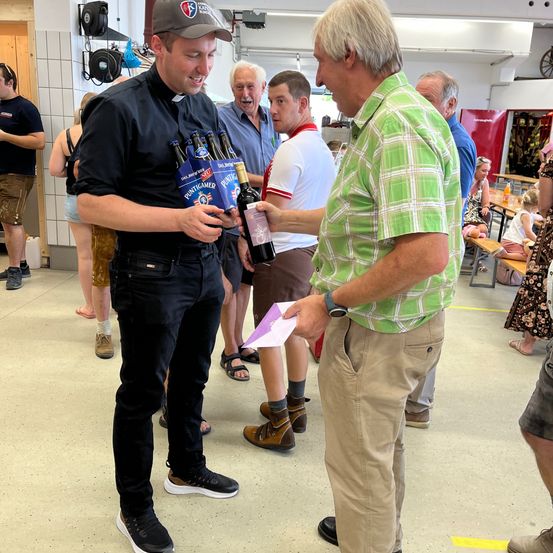Ein Mann in einem schwarzen Hemd und einer Kappe präsentiert eine Schachtel mit Flaschen an einen anderen Mann in einem grünen karierten Hemd. Sie sind von anderen Menschen in einem Raum umgeben.