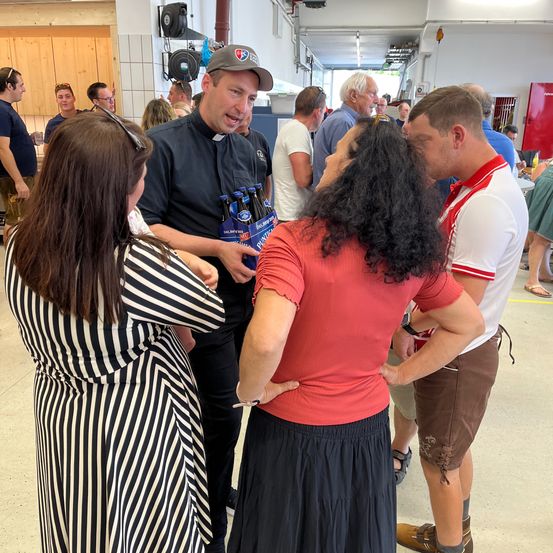 Eine Gruppe von Menschen steht in einem Raum. Ein Mann in einem schwarzen Shirt hält einen Bierkarton. Eine Frau in einem roten Shirt schaut ihn an. Hinter ihnen berührt ein anderer Mann seine Taille.