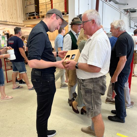 Mehrere Personen stehen in einem Raum, zwei Männer unterhalten sich, während einer eine Schachtel präsentiert. Der Mann in der Kappe schaut sich die Schachtel an. Andere beobachten, einschließlich eines Mannes mit Hut und Brille.
