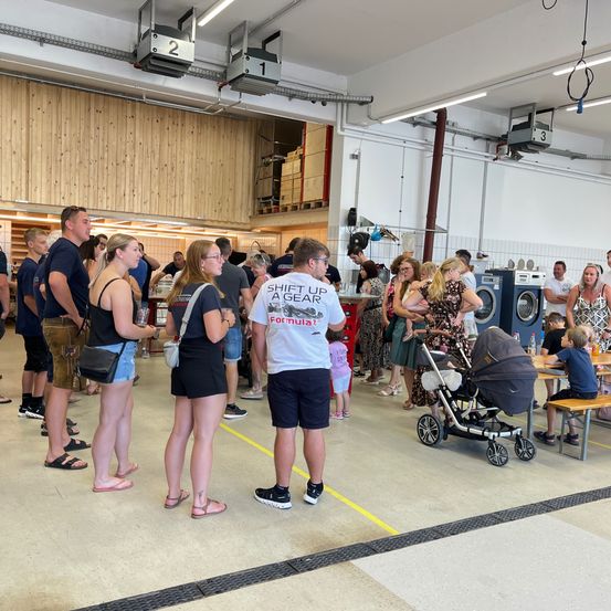 Eine Menschenmenge in einem Raum mit Holzwänden. Einige stehen, andere sitzen. Eine Person mit einem Formel-1-T-Shirt steht in der Mitte. Es gibt einen Kinderwagen, Waschmaschinen und eine Holzbank.