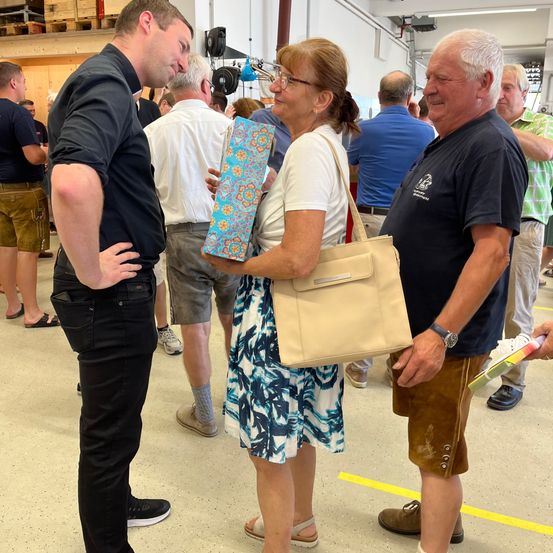 Eine Gruppe von Menschen steht in einem Raum. Eine Frau mit Brille hält ein Geschenk. Vor ihr stehen zwei Männer. Dahinter stehen einige Leute und beobachten sie. Der Boden hat gelbe Markierungen.