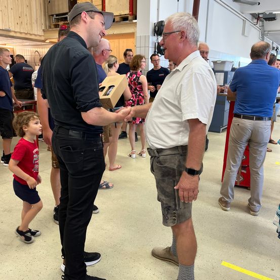 Zwei Männer schütteln sich die Hände in einem Raum mit anderen Leuten. Ein Mann trägt eine Uhr und Lederschuhe. Hinter ihnen stehen und reden Leute. Ein Kind schaut die beiden Männer an.
