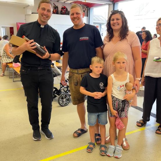 Eine Familie mit zwei Kindern steht in einem Raum mit einem lächelnden Priester, der eine Flasche hält. Hinter ihnen trägt eine Frau ein Baby. Der Raum hat gelbe Markierungen auf dem Boden.