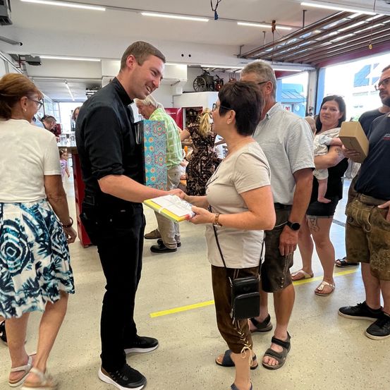 Ein Mann gibt einer Frau ein Papier in einem Raum mit mehreren Personen, einige tragen Kinder. Helle Beleuchtung und ein rollstuhlsichtbar.