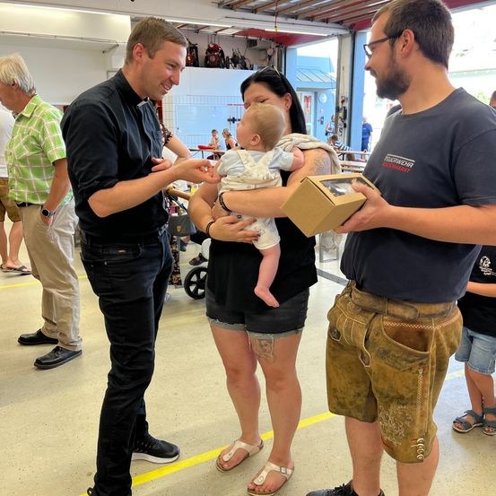 Zwei Männer halten eine Geschenkbox, während eine Frau ein Baby in den Armen hält. Sie stehen in einem Innenraum, möglicherweise einem Gemeindezentrum. Hinter ihnen stehen mehrere Personen und beobachten. Einer von ihnen trägt eine Uhr.