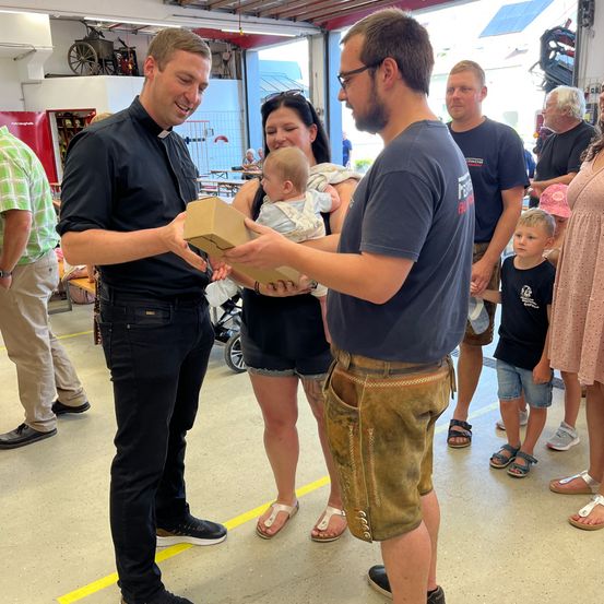 Ein Priester und ein Mann präsentieren einer Frau, die ein Baby hält, eine Box, während mehrere Personen hinter ihnen in einer Feuerwache stehen.