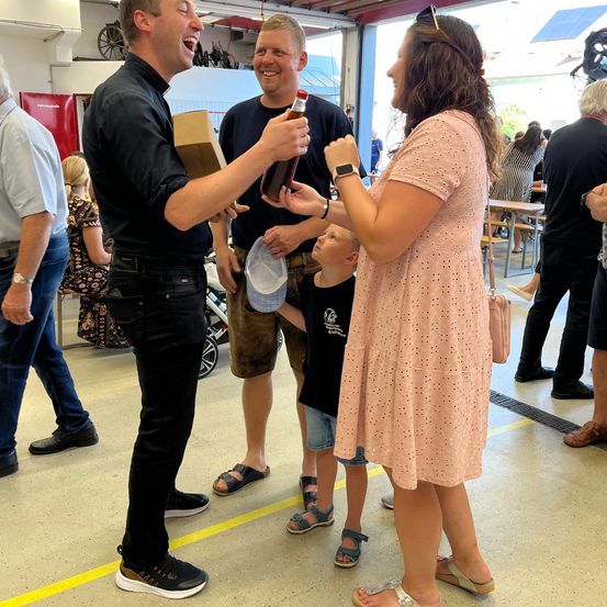 Eine Gruppe von Menschen steht drinnen. Ein Mann reicht einer Frau und einem Jungen eine Flasche. Die Frau und der Junge halten gemeinsam die Flasche. Im Hintergrund sind weitere Menschen zu sehen.