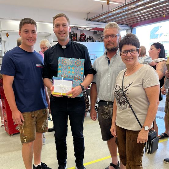 Vier Menschen stehen in einer Werkstatt zusammen. Ein Mann in einem schwarzen Shirt hält ein verpacktes Geschenk. Hinter ihm trägt eine Frau eine Brille und ein Hemd mit Hirschdruck. Der Mann rechts trägt Sandalen und einen Gürtel.