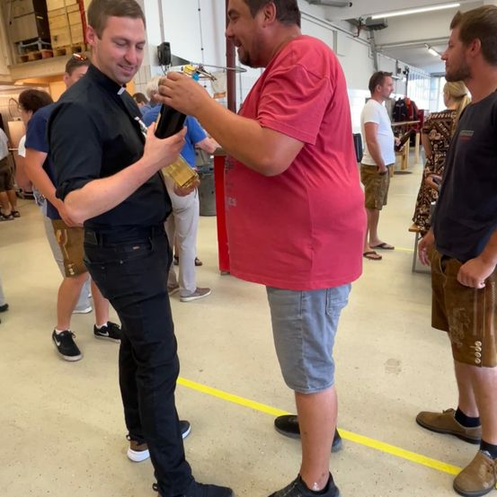 Ein Mann in einem schwarzen Hemd hält eine Flasche und reicht sie einem Mann in einem roten Hemd in einem Raum mit anderen Leuten.