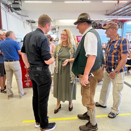 Mehrere Erwachsene unterhalten sich in einem Saal, wobei zwei Männer und eine Frau in der Mitte stehen. Der Mann links trägt ein schwarzes Hemd und eine Hose. Die Frau trägt eine Brille und ein grünes Kleid.