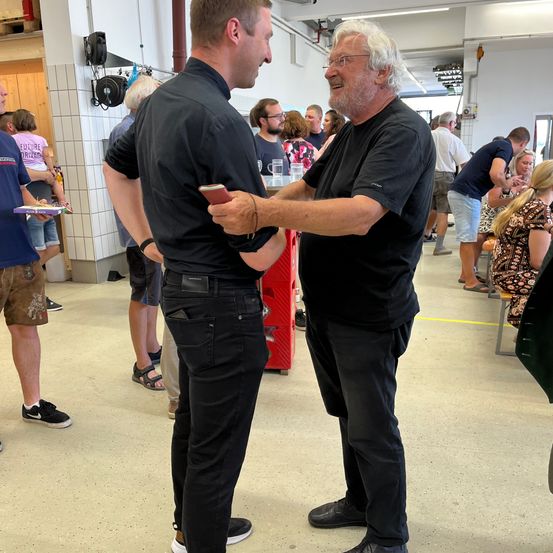 Zwei Männer unterhalten sich in einem überfüllten Raum. Einer hält ein Handy. Andere stehen und sitzen im Hintergrund.