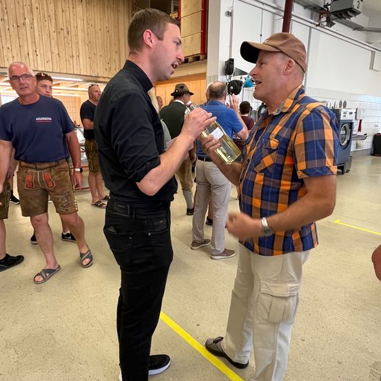 Ein Mann, der eine Flasche Wein in der Hand hält, spricht mit einem anderen Mann in einem Raum mit Holzwänden und einem Fliesenboden. Hinter ihnen stehen andere Menschen und unterhalten sich.
