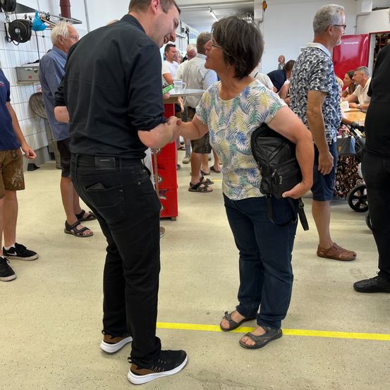 Ein Mann und eine Frau stehen in einer Halle und schütteln Hände. Sie trägt ein Blumenhemd, und er ist in Schwarz. Andere Leute sind um sie herum, einige stehen, andere sitzen an Tischen.