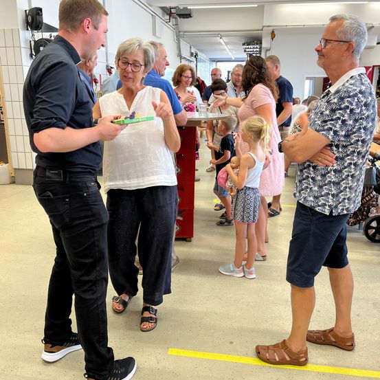 Eine Gruppe von Erwachsenen und Kindern unterhält sich in einem Raum. Ein Mann präsentiert einer älteren Frau etwas, während ein anderer Mann mit verschränkten Armen dahinter steht. Kinder stehen und schauen auf etwas. Der Raum hat weiße Wände und gelbe Markierungen auf dem Boden.
