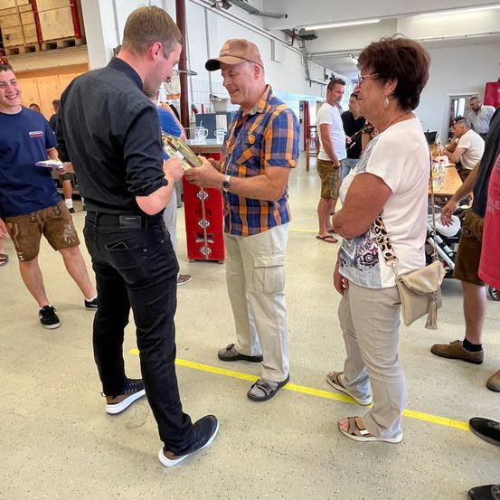 Eine Gruppe von Menschen steht drinnen, möglicherweise bei einer gesellschaftlichen Veranstaltung. Zwei Männer halten eine Trophäe und unterhalten sich. Eine Frau hört in der Nähe zu. Der Raum hat Tische, Stühle und Regale im Hintergrund.