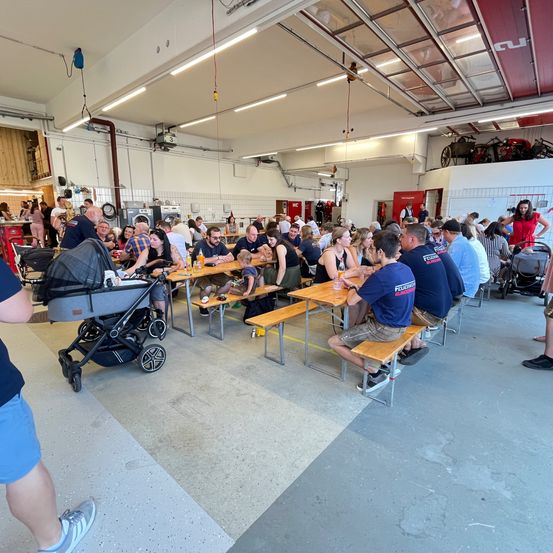 Innenbereich mit vielen Menschen, Tischen und Bänken. Einige tragen Uniformen. Ein Kinderwagen und Fahrräder sind geparkt.