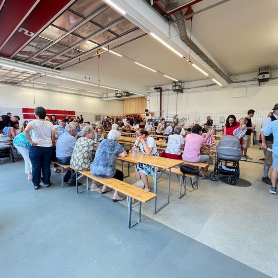 Eine Gruppe von Menschen sitzt an Tischen in einem großen Innenbereich, wahrscheinlich einem Restaurant. Einige tragen Sandalen, andere haben Turnschuhe an. Eines der Kinder ist in einem Kinderwagen.