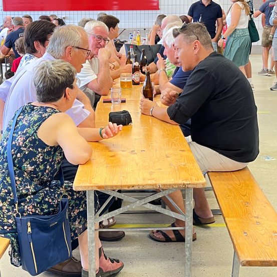 Eine Gruppe von Menschen sitzt an einem langen Holztisch in einem Biergarten. Flaschen und Gläser stehen auf dem Tisch. Ein Mann trägt ein schwarzes Shirt und Sandalen, während andere Freizeitkleidung tragen. Sie führen ein Gespräch.