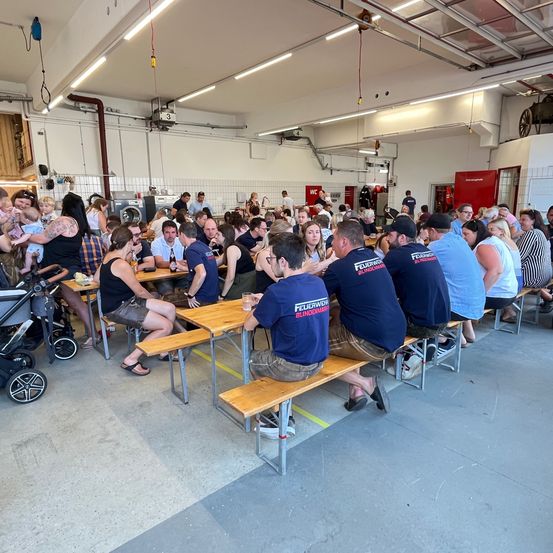 Eine Gruppe von Menschen in blauen Hemden mit 'Feuerwehr' und 'Blindenwerk' Schriftzug sitzen an Tischen in einem Raum mit weißen Wänden. Ein Kinderwagen steht in der Nähe.