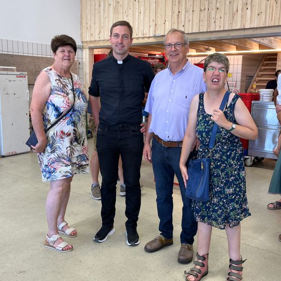 Vier Menschen stehen in einem Gebäude zusammen. Ein Priester in einem schwarzen Gewand steht zwischen einem Mann und einer Frau in Freizeitkleidung. Eine weitere Frau in einem Blumenkleid lächelt. Hinter ihnen steht ein weißer Kühlschrank und eine Treppe.