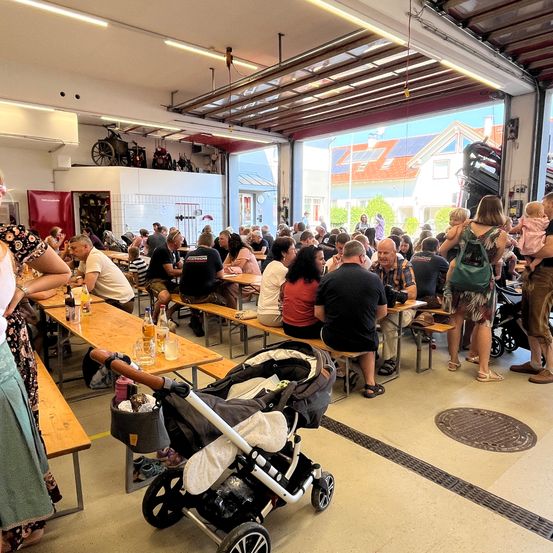 Ein überfüllter Innenbereich mit hölzernen Bänken und Tischen, in dem Menschen verschiedenen Alters sitzen, darunter auch Kinder in Kinderwagen. Der Raum scheint ein umfunktionierter Garagenraum mit hohen Decken und großen Fenstern zu sein.