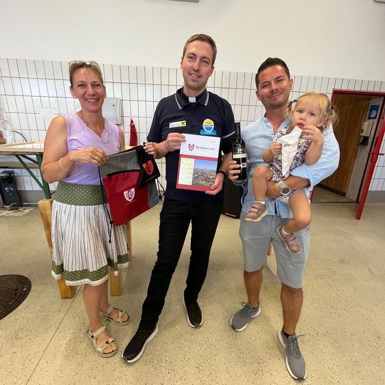 Drei Menschen stehen in einem Raum zusammen. Ein Mann in einem Poloshirt hält ein Buch und eine Flasche. Eine Frau hält eine Tasche, und ein anderer Mann hält ein Baby.