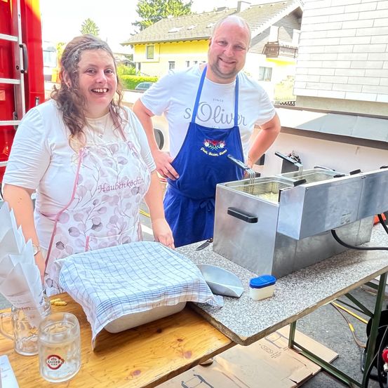 Eine lächelnde Frau und ein Mann stehen neben einem Tisch mit einer Fritteuse, beide tragen Schürzen. Sie scheinen Essen zuzubereiten, wobei im Hintergrund ein Feuerwehrauto zu sehen ist.