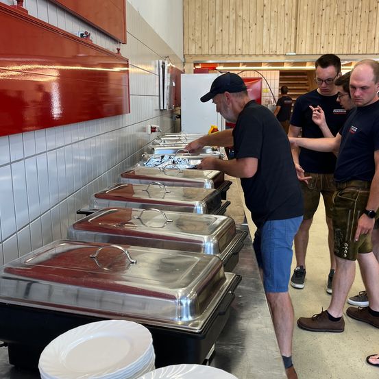 Ein Mann steht an einem Buffettisch mit Essensschalen und Tellern, während sich mehrere andere Personen versammeln. Die Umgebung scheint ein Cafeteria oder Veranstaltungsort zu sein.
