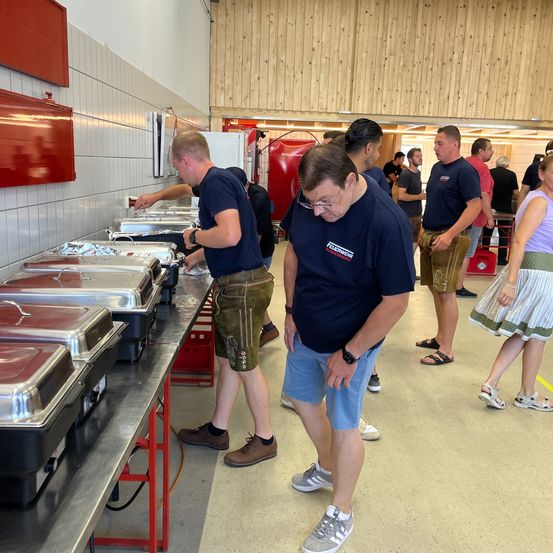 Mehrere Männer und Frauen befinden sich in einem Küchenbereich und bereiten Essen zu. Ein Mann steht an einem Tisch mit Essensbehältern, während ein anderer Mann nach einem Schalter an der Wand greift.