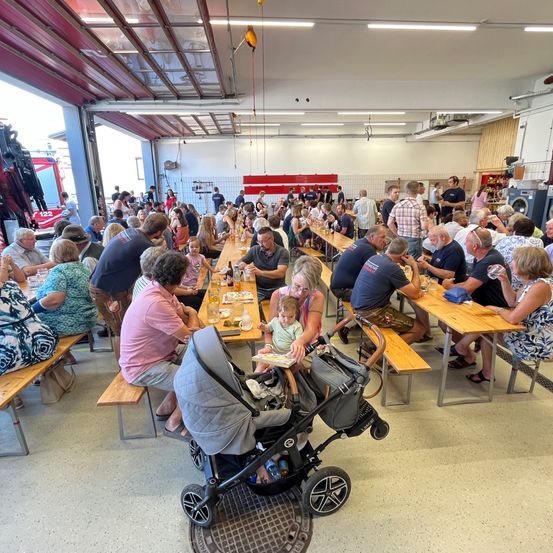 Eine große Gruppe von Menschen versammelt sich in einem Raum mit Holztischen und -bänken. Sie sitzen und stehen und unterhalten sich. Einige essen und trinken. Ein Baby sitzt auf dem Schoß einer Frau.