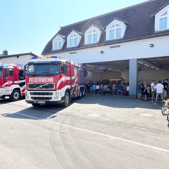 Ein roter Feuerwehrwagen steht vor einem Gebäude, in dessen Nähe sich mehrere Menschen versammelt haben. Das Gebäude hat mehrere Fenster und ein Dach mit Lampen. Ein weiterer Feuerwehrwagen ist links geparkt.