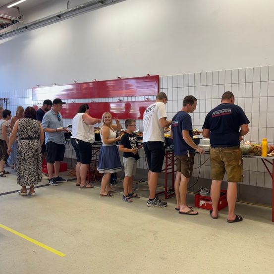 In einer Feuerwache stehen mehrere Menschen in einer Schlange, um zu essen, wahrscheinlich an einem Buffet. Eine Frau hält einen kleinen Jungen an der Hand.