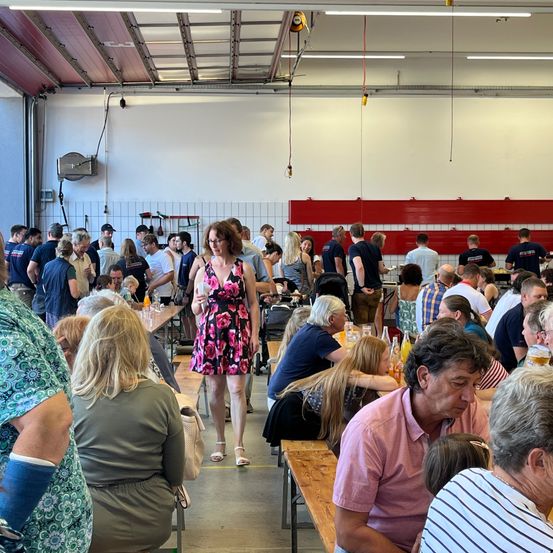 Ein großer Innenbereich zum Essen mit vielen Menschen an Tischen. Eine Frau in einem Blumenkleid geht zwischen den Tischen hindurch. Der Raum hat weiße Wände, rote Akzente und Deckenbeleuchtung.