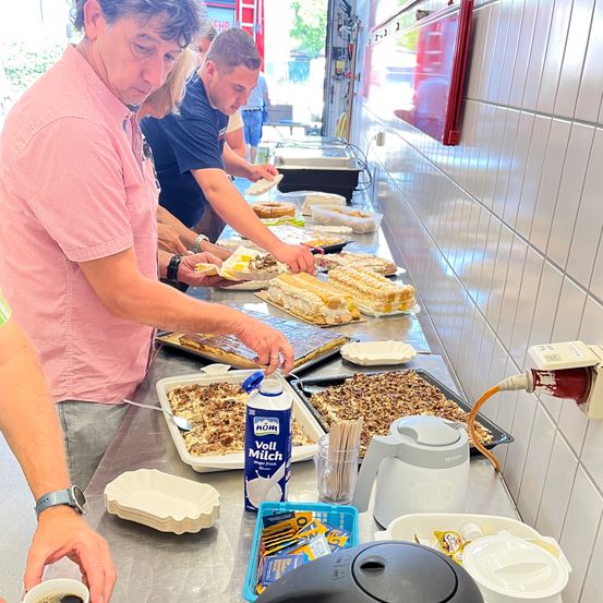 Mehrere Personen bereiten Essen an einem Tisch mit Desserttabletts, einer Milchflasche und einer Kaffeemaschine vor. Der Tisch ist mit Tassen, Tellern und anderen Utensilien bestückt.