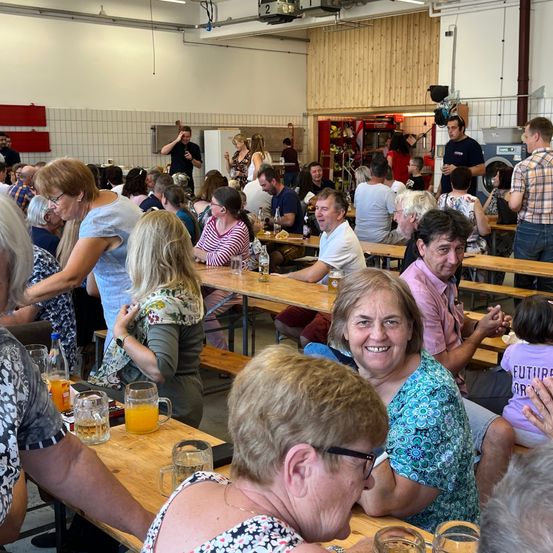 Ein Innenrestaurant mit Holztischen und Bänken. Viele Menschen sitzen und stehen, einige lächeln und es gibt Getränke auf den Tischen. Ein Mann in einem blauen Hemd spricht am Telefon.