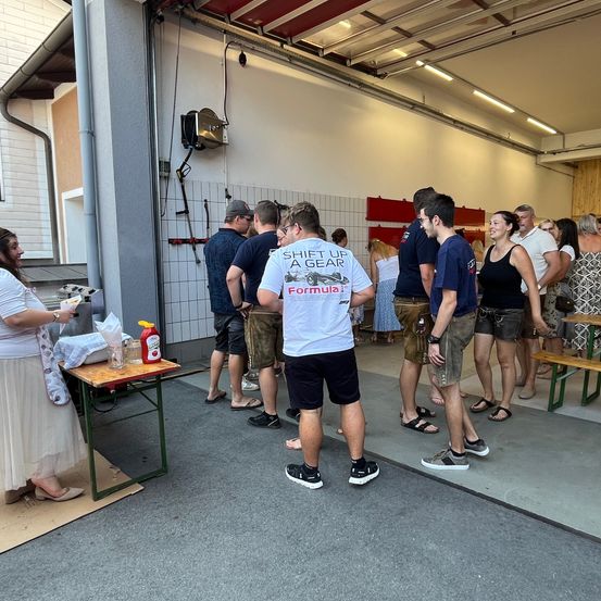 Eine Gruppe von Menschen in einer Garage, einer trägt ein weißes T-Shirt mit der Aufschrift 'Shift Up A Gear Formula 1', möglicherweise zur Vorbereitung auf eine Veranstaltung.