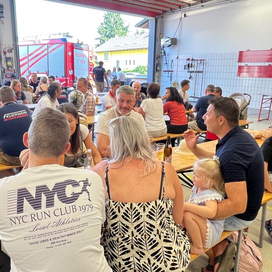 Eine Gruppe von Menschen sitzt in einer Feuerwache um Tische herum. Eine Frau in einem schwarz-weißen Kleid schaut einen lächelnden Mann an. Ein Mann hält ein Mädchen auf seinem Schoß. Ein Feuerwehrwagen ist draußen geparkt.