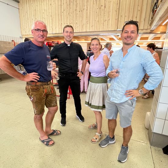 Vier Personen stehen in einem Innenraum und lächeln für ein Foto. Ein Mann auf der linken Seite hält einen Becher, während ein anderer Mann neben ihm eine Uhr trägt. Die Frau trägt einen Rock und Sandalen. Der Boden ist gefliest und hinter ihnen befindet sich eine Holzwand.