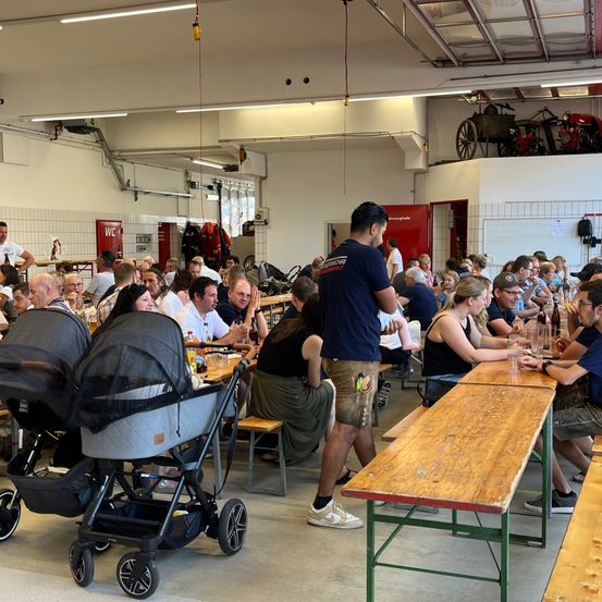 Ein überfüllter Speisebereich mit mehreren Menschen, die an Tischen sitzen. Ein Mann geht auf einen Tisch mit einem Baby zu. Im Hintergrund ist ein Feuerwehrwagen ausgestellt.