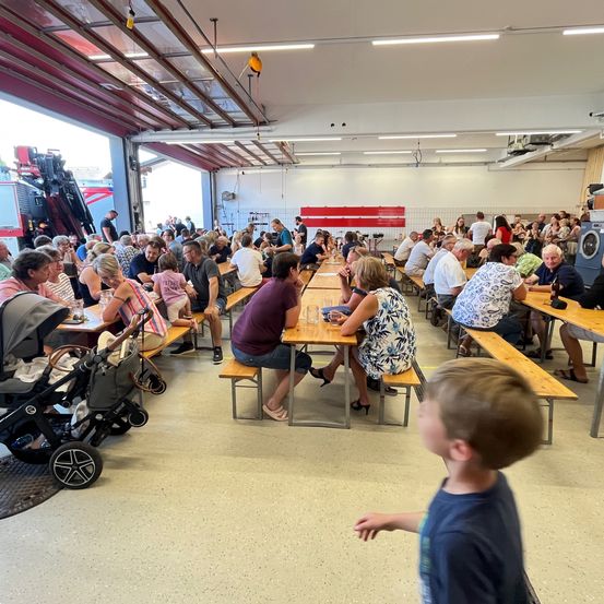 Ein großer Raum mit Tischen und Bänken, gefüllt mit Menschen, möglicherweise für ein gemeinschaftliches Treffen. Ein Feuerwehrauto ist auf der linken Seite geparkt.