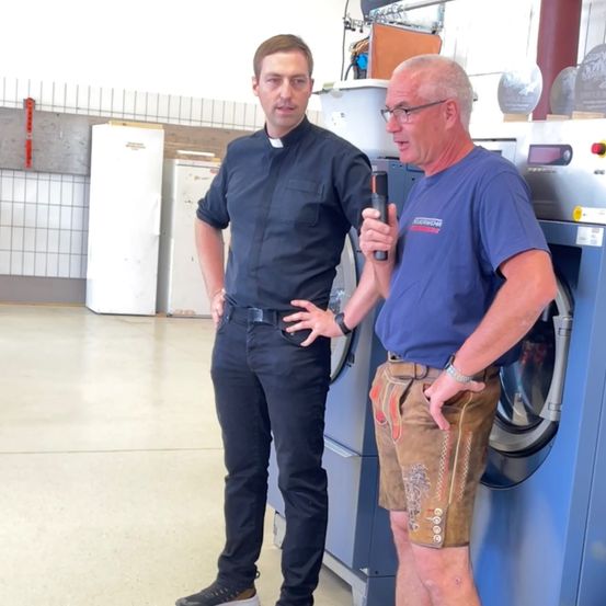 Zwei Männer stehen vor Waschmaschinen, einer hält ein Mikrofon, gekleidet in einem Hemd und Shorts, der andere in einem Hemd und Hose. Hinter ihnen befinden sich Kühlschränke und geflieste Wände.