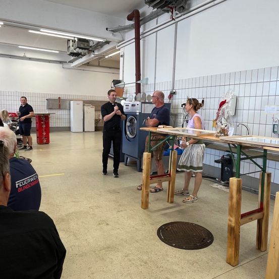 Mehrere Personen versammeln sich in einem Raum mit gefliesten Wänden, ein Mann spricht in ein Mikrofon, während andere in der Nähe einer Waschmaschine, eines Tisches und einer Tür stehen.