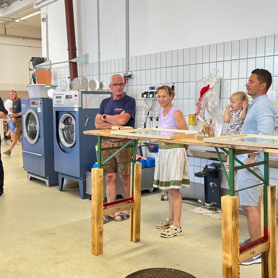 Eine Gruppe von Menschen befindet sich in einer Fabrik, wobei ein Mann und eine Frau in der Nähe eines Tisches stehen, während ein Baby auf dem Tisch steht. Dahinter stehen zwei Waschmaschinen.