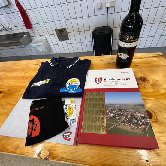 Ein Holztisch hat ein Buch, eine Flasche Wein, ein Polohemd und einen schwarzen Stoff mit Text darauf. Dahinter befinden sich ein Waschbecken, ein Mülleimer und ein Lautsprecher.