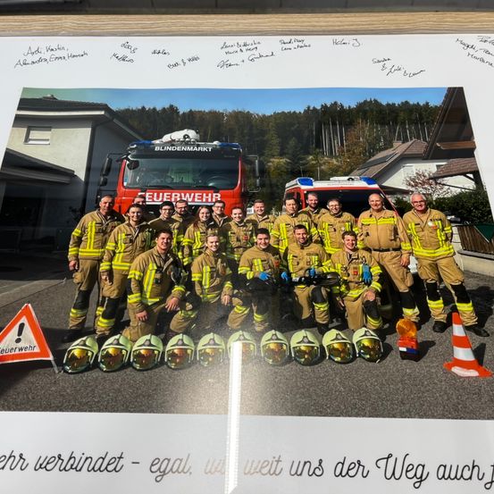 Ein Foto von Feuerwehrleuten in Uniform, die vor einem Feuerwehrwagen lächelnd und posierend fotografiert wurden. Oben befinden sich Unterschriften und unten eine Botschaft.